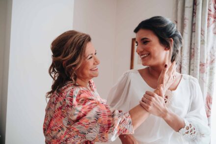 La novia, ya vestida, se ríe y sujeta junto al pecho la mano de una invitada de blusa estampada