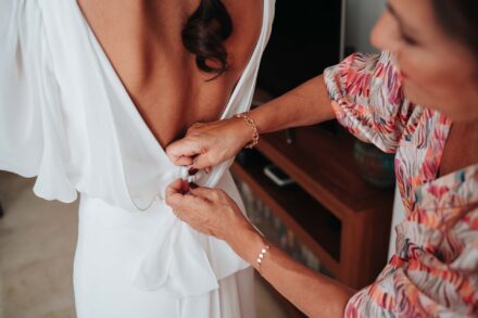 Una invitada de blusa estampada abrocha el botón del escote en la espalda del vestido de la novia
