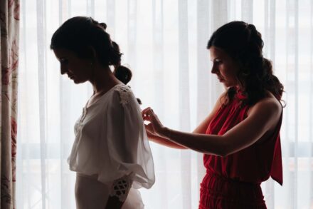 Una invitada de vestido rojo abrocha la espalda del vestido de la novia a contraluz de la ventana