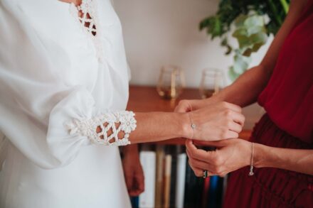 Una invitada de vestido rojo le abrocha a la novia una pulsera fina bajo el puño de encaje