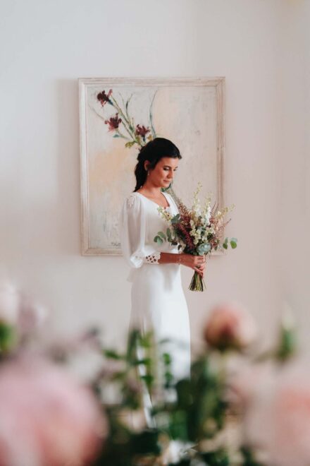La novia, de pie ante un cuadro, mira su ramo de flores blancas y granates, entre flores desenfocadas