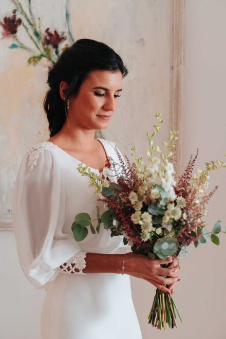 La novia, de cerca, sonríe mirando su ramo con eucalipto, espigas granates y flores blancas