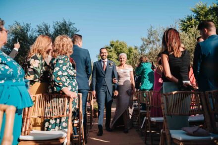 El novio sonríe al entrar a la ceremonia del brazo de una mujer de pelo cano con vestido gris