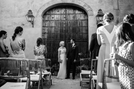 El novio y su acompañante de vestido claro esperan ante el portón de madera, en blanco y negro