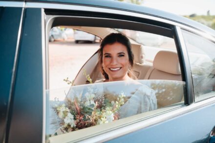 La novia sonríe desde el asiento trasero del coche, con el ramo de flores blancas y granates junto a la ventanilla
