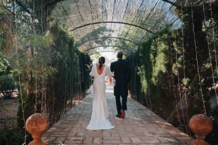 De espaldas, la novia y un hombre de chaqué avanzan bajo un túnel de cañizo entre cipreses