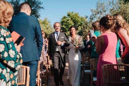 La novia avanza sonriente por el pasillo de sillas del brazo de un hombre de chaqué gris, con el ramo
