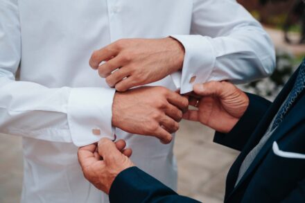 Un hombre de traje azul marino le pone los gemelos al novio en los puños de la camisa blanca