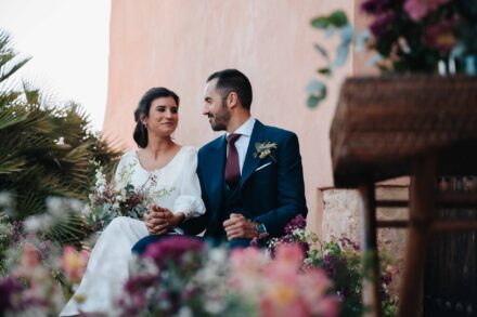 Sentados en la ceremonia, el novio mira a la novia, que sonríe, tras un primer plano de flores rosas y moradas