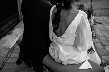 En blanco y negro, la novia sentada de espaldas junto al novio, con escote en V, botones y lazo en el vestido