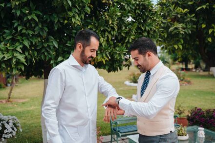Un invitado con chaleco beis y corbata azul le abrocha el reloj al novio en el jardín