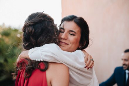 La novia, con los ojos cerrados, abraza con fuerza a una invitada de vestido rojo