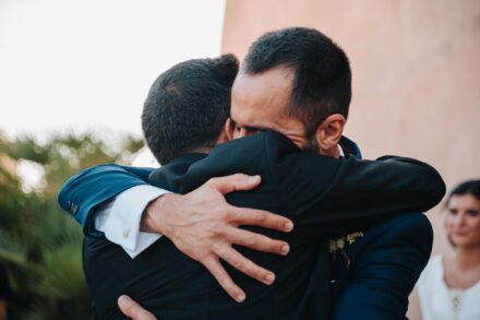 El novio abraza con los ojos cerrados a un invitado de chaqué mientras la novia mira desde el fondo