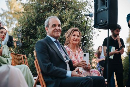 Un invitado de traje gris y una invitada con el móvil en la mano sonríen sentados durante la ceremonia