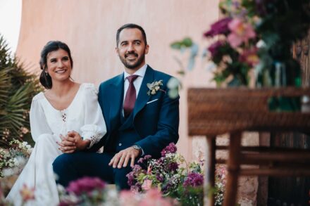 Los novios, sentados y de la mano, sonríen mirando hacia los invitados durante la ceremonia civil