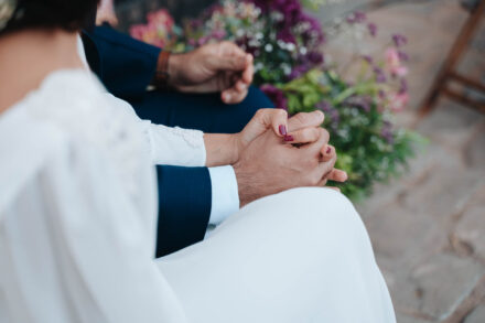 Las manos entrelazadas de los novios sentados en la ceremonia, con las uñas granates de la novia