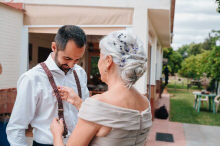 Una invitada de pelo canoso con flores en el recogido le coloca los tirantes al novio, que sonríe