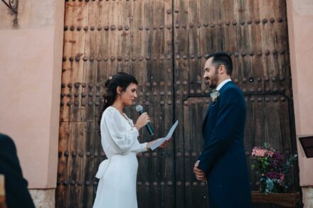 La novia lee un papel al micrófono frente al novio, ante el gran portón claveteado de madera