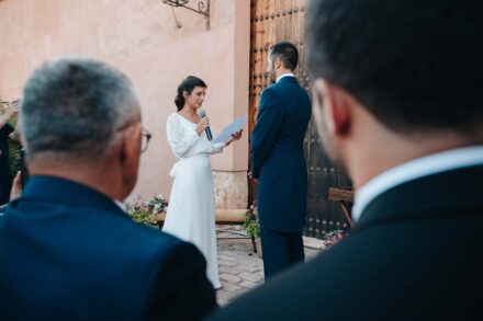 Entre las cabezas de dos invitados, la novia lee con el micrófono frente al novio