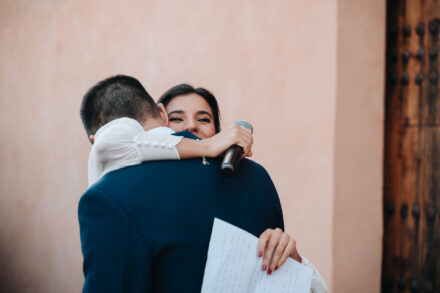 La novia abraza al novio con el micrófono y la hoja todavía en las manos, mirando a cámara