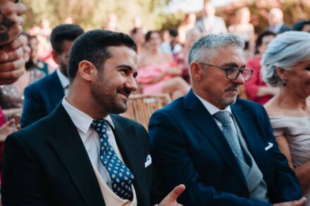 Un invitado joven con corbata de lunares sonríe junto a otro de gafas, sentados en la ceremonia