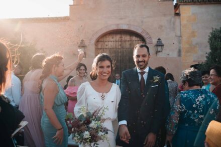 Los novios salen de la ceremonia de la mano bajo una lluvia de pétalos, a contraluz