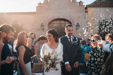 La novia ríe al salir de la mano del novio mientras los invitados les lanzan confeti blanco
