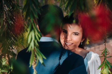 La novia sonríe a cámara abrazada al novio, vista entre ramas colgantes con flores rojas