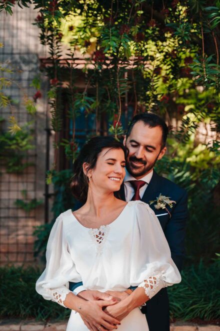 El novio abraza por la espalda a la novia, que sonríe con los ojos cerrados bajo una enredadera