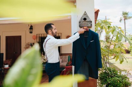 El novio, con tirantes, cuelga su chaqué azul de la casita de pájaros del porche
