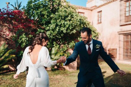 Los novios bailan cogidos de la mano en el césped, con la novia de espaldas, frente a la fachada rosada de la hacienda