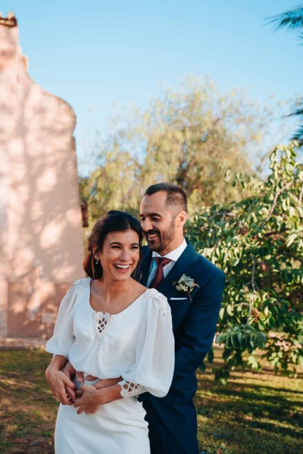 El novio abraza por detrás a la novia, que se ríe a carcajadas junto a un muro rosado al sol