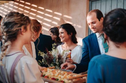 La novia sonríe con una cerveza y el ramo mientras un invitado coge queso de una bandeja en el cóctel