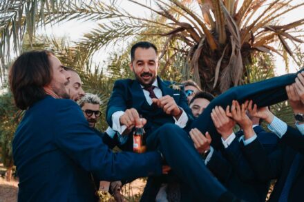 Varios invitados levantan en volandas al novio, que sonríe con una botella en la mano bajo una palmera