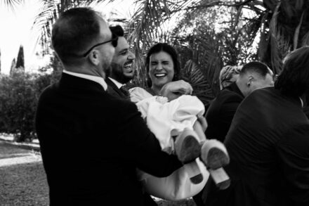 Varios invitados cogen en brazos a la novia, que se ríe con los ojos entornados, en blanco y negro