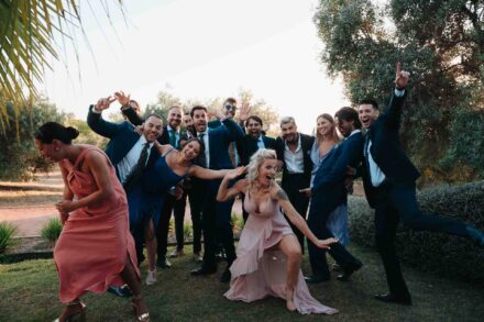 Un grupo de invitados posa entre gritos y gestos exagerados, con una invitada de rosa agachada delante