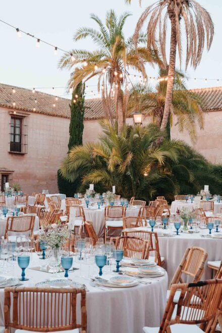 Mesas del banquete con copas azules y sillas de bambú en el patio, bajo palmeras y guirnaldas de luces