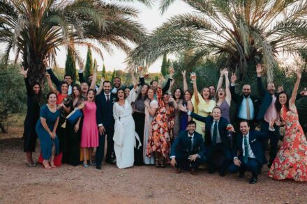 Los novios posan con un grupo grande de invitados que levantan brazos y copas bajo las palmeras