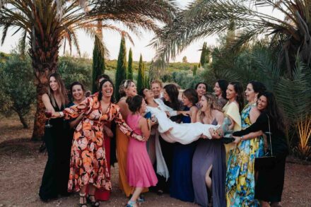 Un grupo de invitadas sostiene en brazos a la novia, tumbada y riendo, bajo las palmeras