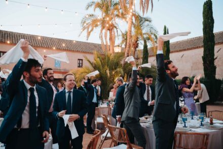 Los invitados, de pie junto a las mesas del banquete, agitan servilletas blancas bajo las guirnaldas de luces