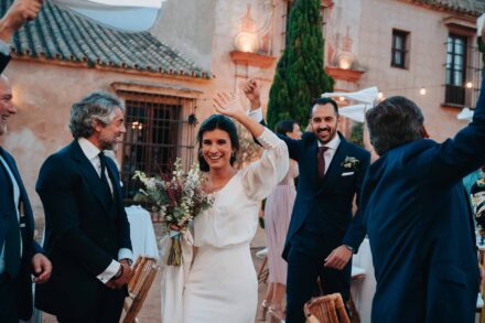 La novia, con el ramo, y el novio entran al banquete saludando con la mano en alto entre los invitados
