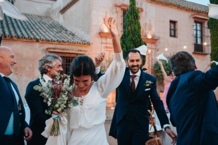 La novia se ríe mirando al suelo con el brazo en alto, seguida del novio, entre invitados que agitan servilletas