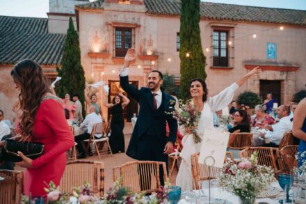 Los novios saludan con los brazos en alto entre las mesas del banquete, junto al cartel de la mesa 1
