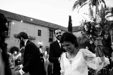 La novia levanta el ramo y sonríe con la mirada baja mientras el novio, detrás, se ríe, en blanco y negro