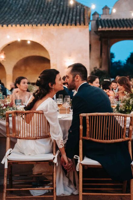 Los novios, sentados de espaldas en la cena y cogidos de la mano, se miran bajo los arcos iluminados
