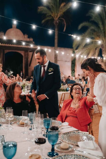 En el banquete, la novia ríe junto a una invitada mayor vestida de rojo y el novio charla con otra en la mesa