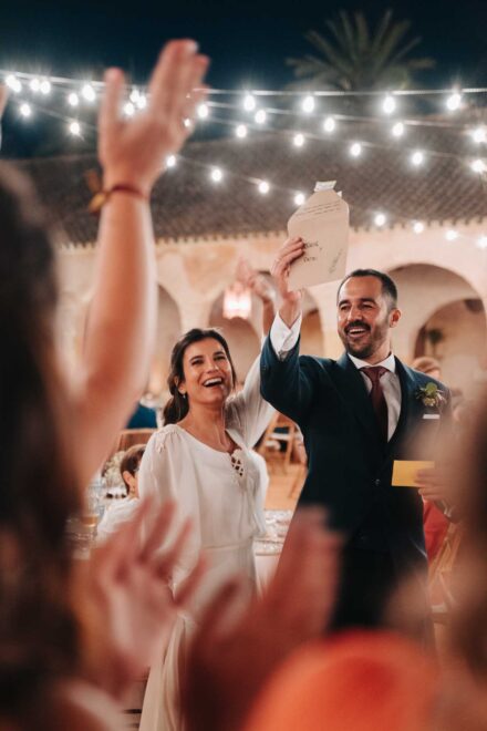 Los novios ríen y el novio levanta un sobre escrito a mano mientras los invitados alzan las manos