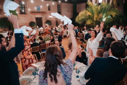 Los invitados agitan servilletas en el aire y la novia levanta el brazo entre las mesas del banquete