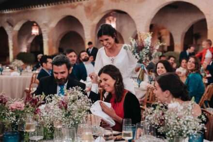 La novia, con el ramo en la mano, se acerca sonriendo a una mesa de invitados bajo los arcos del patio