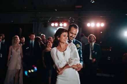 El novio abraza a la novia por detrás mientras bailan en la pista bajo los focos, ante los invitados